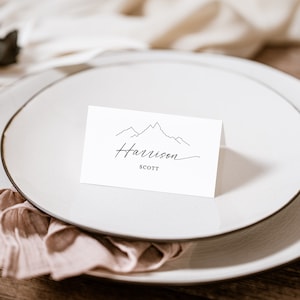 May include: White place card with a simple line drawing of mountains and the text "Harrison SCOTT" in black cursive.