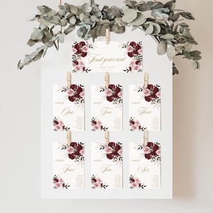 May include: A wedding seating chart display featuring a white board with seven rectangular cards attached by wooden clips. The cards are decorated with burgundy and dusty pink floral illustrations and gold text, including Find your seat.