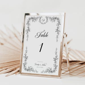 May include: A silver frame with a white card inside. The card has a floral border and the text "Table 1" in black. The text "Joseph & Julian 08.06.20XX" is also printed on the card.