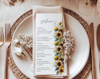 May include: A formal table setting with a white plate, gold cutlery, and a menu card. The menu card features a floral design of yellow sunflowers and the word "Menu" in elegant script. The setting includes a woven placemat and a folded napkin.