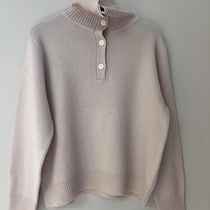 May include: A light beige sweater with a high, ribbed collar and three large, round buttons down the front. The sweater has long sleeves and a ribbed hem. It is hanging on a metal hanger against a plain background.