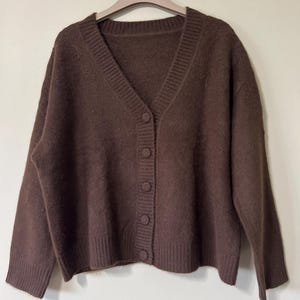 Puede incluir: Un cárdigan marrón con cuello en V y cierre de botones. El suéter está hecho de un material suave y texturizado y tiene puños y dobladillo acanalados. Los botones son del mismo color que el suéter. Cuelga de una percha beige.
