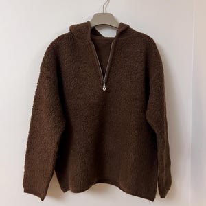 Pode incluir: Um suéter marrom texturizado com fecho de meio zíper e capuz. O suéter está pendurado em um cabide contra uma parede branca. O material parece macio e felpudo.
