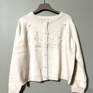 以下が含まれることがあります： 花柄の刺繍とボタン留めが施されたクリーム色のカーディガン。ニュートラルな背景にハンガーにかけられています。丸首のセーターは長袖で、リブ編みの裾が付いています。