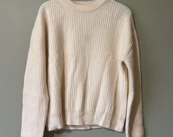 Könnte beinhalten: Cremefarbener Rippstrickpullover mit Rundhalsausschnitt und langen Ärmeln. Der Pullover hängt an einem grauen Kleiderbügel vor einem gedämpften grünen Hintergrund. Das Strickmuster ist vertikal und gleichmäßig.
