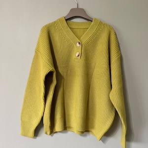 May include: A mustard yellow knitted sweater with a V-neck and long sleeves. The sweater has a ribbed texture and two gold-coloured buttons at the neckline. It is hanging on a wooden hanger against a neutral background.