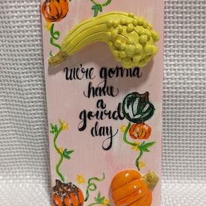 May include: A pink wooden sign with the text "We're gonna have a gourd day" painted in black. The sign is decorated with painted vines, yellow flowers, and three-dimensional gourds and pumpkins.