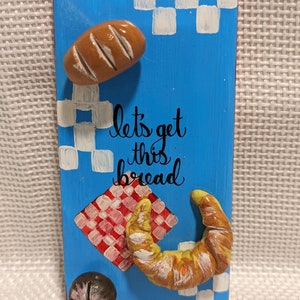 Puede incluir: Un letrero de madera azul con el texto "let's get this bread" pintado en negro. El letrero tiene un patrón de cuadros rojos y blancos, una hogaza de pan marrón, un croissant y un botón blanco y negro.