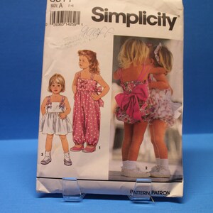 Puede incluir: Patrón de costura Simplicity número 8511, talla A (1-4), para vestidos y un mono para niñas. El patrón muestra tres estilos diferentes: un vestido blanco con un lazo, un mono rosa de lunares con un lazo y un vestido rosa floral con un gran lazo rosa.