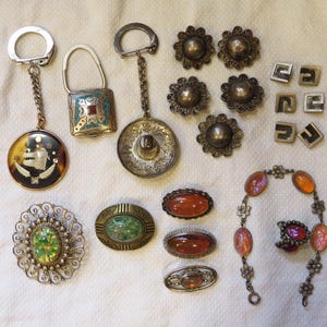 May include: A collection of vintage jewelry and accessories, including keychains, brooches, earrings, and a bracelet. The items feature various designs, colors, and materials, such as silver, turquoise, and amber-colored stones. The items are displayed on a white surface.