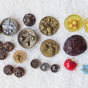 May include: A collection of vintage buttons in various sizes and designs. The buttons feature fruit and floral motifs, including cherries, pears, and grapes. Colors include gold, silver, brown, red, and yellow. A wooden ruler is on the left.