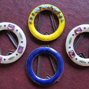 May include: Four decorative metal picture frames with enamel accents. The frames are circular and feature a variety of colors and designs, including yellow with blue flowers, white with purple gems, and blue with a white border.