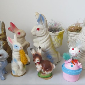 May include: A collection of vintage Easter bunny figurines in various poses and colors. The bunnies are painted in pastel shades of blue, pink, and gold, with red eyes and some holding carrots. Some bunnies have baskets on their backs.