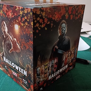 Op de afbeelding: Een Halloween filmcollectie boxset met Michael Myers. De doos heeft afbeeldingen van de gemaskerde moordenaar en de filmtitel. De boxset staat op een groen oppervlak met een pen en een schaar.