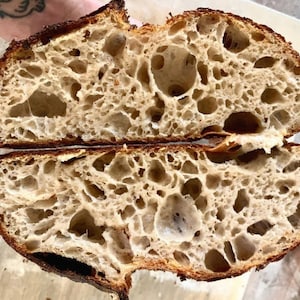 May include: A close-up of a sliced loaf of bread with a light brown crust and a very airy, open crumb. The bread has a slightly irregular shape and is sliced in half, revealing the interior.