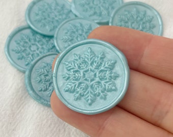 May include: A close-up of a light blue wax seal with a snowflake design. The seal is being held by a hand.