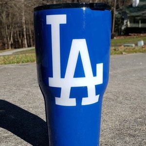 May include: A blue and white tumbler with the LA logo. The tumbler is made of stainless steel and has a black lid.