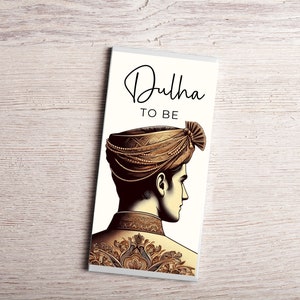 May include: A white chocolate bar featuring a gold illustration of a man in a turban and traditional Indian attire. The text "Dulha to Be" is printed on the bar.
