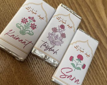 Puede incluir: Tres pequeñas barras de chocolate envueltas en papel de aluminio plateado con etiquetas blancas. Cada etiqueta presenta un diseño floral diferente y el texto "Eid Mubarak" en escritura árabe. Los nombres "Krinnt", "Tryfonh" y "Sann" están escritos en las etiquetas.