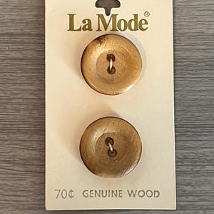 May include: Two round, light brown wooden buttons from La Mode, attached to a cream-colored card. The card reads "70¢ GENUINE WOOD" in black text. The buttons have a natural wood grain and a four-hole design.