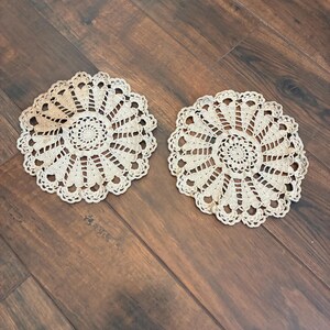 May include: Two beige, circular crochet doilies with a detailed, openwork design. The doilies are placed on a dark brown wooden surface. The crochet pattern features a central spiral surrounded by radiating lines and decorative edging.