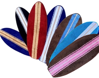 May include: A collection of seven surfboard-shaped rugs arranged in a fan pattern. Each rug features a solid color base including red, blue, black, brown, and turquoise, with a contrasting vertical stripe running down the center of each piece.