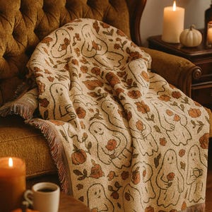 Peut inclure: Couverture beige avec un motif répété de fantômes amicaux et de fleurs orange. La couverture est drapée sur un fauteuil marron. La couverture a une bordure à franges et est décorée sur le thème d'Halloween.