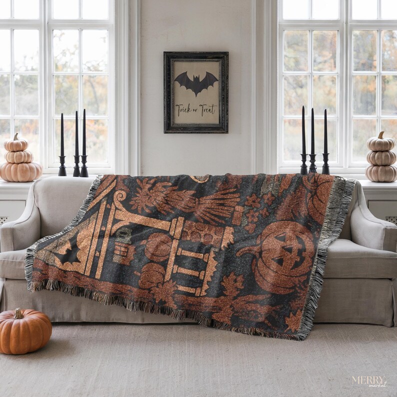 May include: A Halloween-themed throw blanket with a black and orange design featuring pumpkins, bats, and other festive elements. The blanket is draped over a beige sofa, with a framed picture and candles in the background.