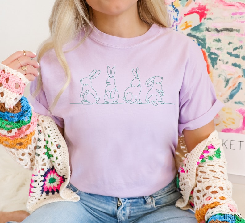 May include: A lavender t-shirt with a line drawing of four bunnies on the front. The bunnies are facing to the right and are in a line.