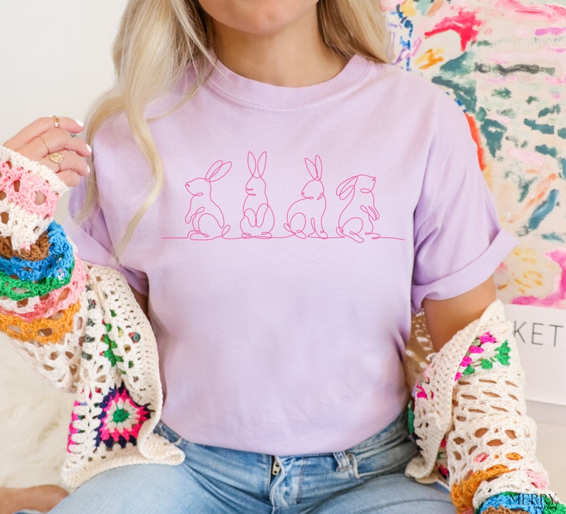 May include: A light purple t-shirt with a line drawing of five bunnies in pink. The bunnies are facing forward and are in a row. The t-shirt is being worn by a person wearing blue jeans.
