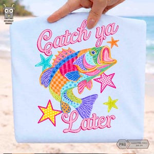 May include: Light blue t-shirt featuring a colorful fish illustration with the words "Catch ya Later" in pink script. The fish is decorated with various patterns and colors, surrounded by star shapes.