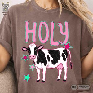 Peut inclure: T-shirt marron avec le mot "HOLY" en rose bord&eacute; de vert. En dessous, une vache noire et blanche et des &eacute;toiles roses. Le t-shirt est en mati&egrave;re souple.