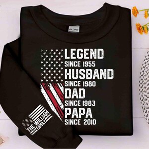 Puede incluir: Sudadera negra con un dise&ntilde;o de bandera estadounidense y el texto "LEGEND SINCE 1955, HUSBAND SINCE 1980, DAD SINCE 1983, PAPA SINCE 2010." La manga muestra "THE WILLIAM FAMILY" y una peque&ntilde;a bandera.
