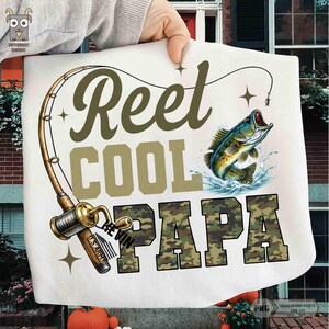 May include: White t-shirt with a fishing theme. The design includes a fishing rod, a leaping fish, and the words "Reel Cool Papa" in olive green and camouflage lettering. The shirt has a graphic design.