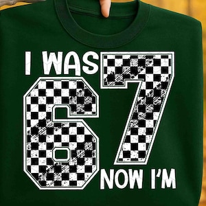 May include: Dark green sweatshirt with the text "I WAS 67 NOW I'M" in white. The numbers 6 and 7 are designed with a black and white checkered pattern. The sweatshirt is a crew neck style.