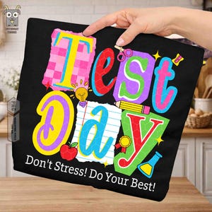May include: Black t-shirt with the words "Test Day" in colorful, block letters. The design includes a lightbulb, pencil, apple, and other school-themed graphics. The text "Don't Stress! Do Your Best!" is printed below.