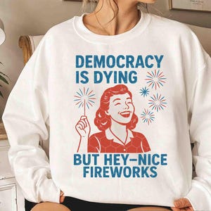 May include: White sweatshirt with the text "DEMOCRACY IS DYING BUT HEY-NICE FIREWORKS" in blue. The graphic features a smiling woman with red hair and a red and white outfit holding a sparkler. The design includes red and blue fireworks.