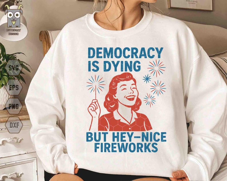 May include: White sweatshirt with the text "DEMOCRACY IS DYING BUT HEY-NICE FIREWORKS" in blue. The graphic features a smiling woman with red hair and a red and white outfit holding a sparkler. The design includes red and blue fireworks.