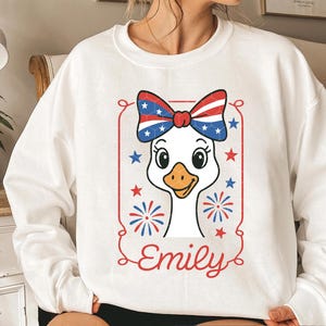 May include: White sweatshirt with a red, white, and blue goose wearing a bow. The goose has a white body and a red beak. The bow is red, white, and blue with stars. The text "Emily" is below the goose.