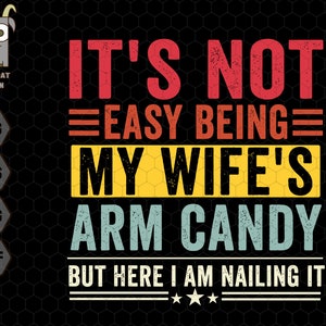 May include: A vintage-style graphic with the text "It's not easy being my wife's arm candy but here I am nailing it". The text is in red, yellow, blue, and white with a black background.