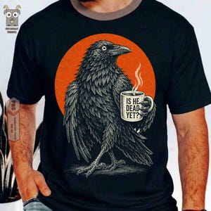 Könnte beinhalten: Schwarzes T-Shirt mit einer detaillierten Illustration eines Raben, der eine Kaffeetasse hält. Der Rabe ist vor einem orangefarbenen Kreis mit Wabenmuster platziert. Auf der Tasse steht "IS HE DEAD YET?"