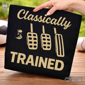Könnte beinhalten: Schwarzes T-Shirt mit goldenem Text "Classically TRAINED". Das Design zeigt Illustrationen von Autopedalen und einem Schalthebel. Das T-Shirt wird von einer Hand gehalten.
