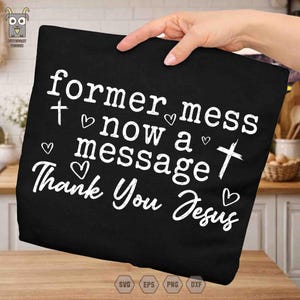 Könnte beinhalten: Schwarzes T-Shirt mit weißem Text: "former mess now a message Thank You Jesus" mit Kreuz- und Herzsymbolen. Das T-Shirt wird von einer Hand gehalten.