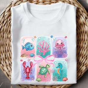 May include: A white t-shirt with a colorful design featuring a blue fish, a pink jellyfish, a red lobster, a green sea turtle, and a pink coral. The design is surrounded by a pink ribbon.