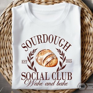 Op de afbeelding: Wit T-shirt met een bruin grafisch ontwerp met de tekst "Sourdough Social Club Wake and Bake" en een tekening van een zuurdesembrood. De tekst "Est 2025" is ook in het ontwerp opgenomen.