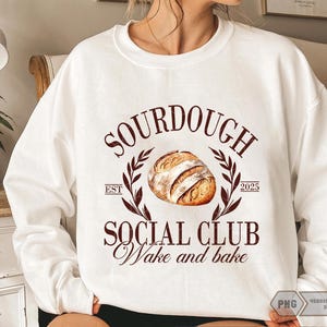 Op de afbeelding: Wit sweatshirt met een bruine afbeelding waarop "Sourdough Social Club Wake and Bake" staat, met een illustratie van een zuurdesembrood en de tekst "Est. 2025".
