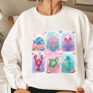 May include: White sweatshirt with a colorful graphic print of a seahorse, a turtle, a lobster, a jellyfish, a fish, and seaweed. The graphic is surrounded by a pink ribbon and blue and white bubbles.