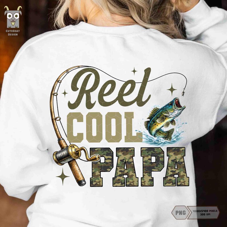 May include: White sweatshirt with a fishing-themed graphic. The design features the words "Reel Cool Papa" in green and camouflage lettering, a fishing rod, and a leaping fish. The sweatshirt has a crew neck.