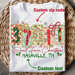 Könnte beinhalten: Weißes T-Shirt mit einem weihnachtlichen Design. Das Design beinhaltet die Zahlen "37011" in festlichen Mustern, den Text "Small Town Christmas" und "NASHVILLE, TN". Das T-Shirt hat einen Rundhalsausschnitt und wird auf einer gewebten Oberfläche präsentiert.
