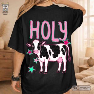Peut inclure: T-shirt noir avec un motif de vache et le mot "HOLY" en rose. La vache a des taches noires et blanches, avec des &eacute;toiles roses et turquoise. Le t-shirt a des manches courtes.